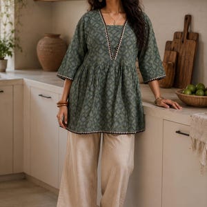 May include: A woman wearing a green patterned tunic top with a square neckline and three-quarter sleeves, paired with beige wide-leg pants. The top has a decorative trim. The outfit is casual and comfortable.