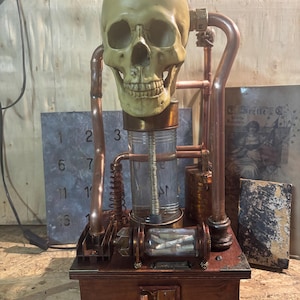 May include: A decorative sculpture featuring a beige human skull mounted atop a clear glass cylinder and a dark wooden base. The structure is accented with copper pipes, a small horizontal glass vial containing metal pieces, and metal corner brackets.