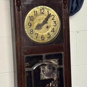 May include: A dark brown antique wall clock with a gold-coloured clock face. The clock has a rectangular wooden frame with decorative carvings and a mirrored lower section. The clock face displays Roman numerals.