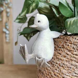 May include: A white ceramic duck ornament perched on a woven basket. The duck has a detailed beak and webbed feet, with its wings slightly outstretched. Green leafy plants are in the background, adding a natural touch to the scene.
