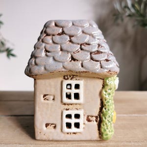 May include: A ceramic house figurine with a gray, textured roof and a beige body. The house has two square windows and a green vine detail on the side. The figurine is on a wooden surface.