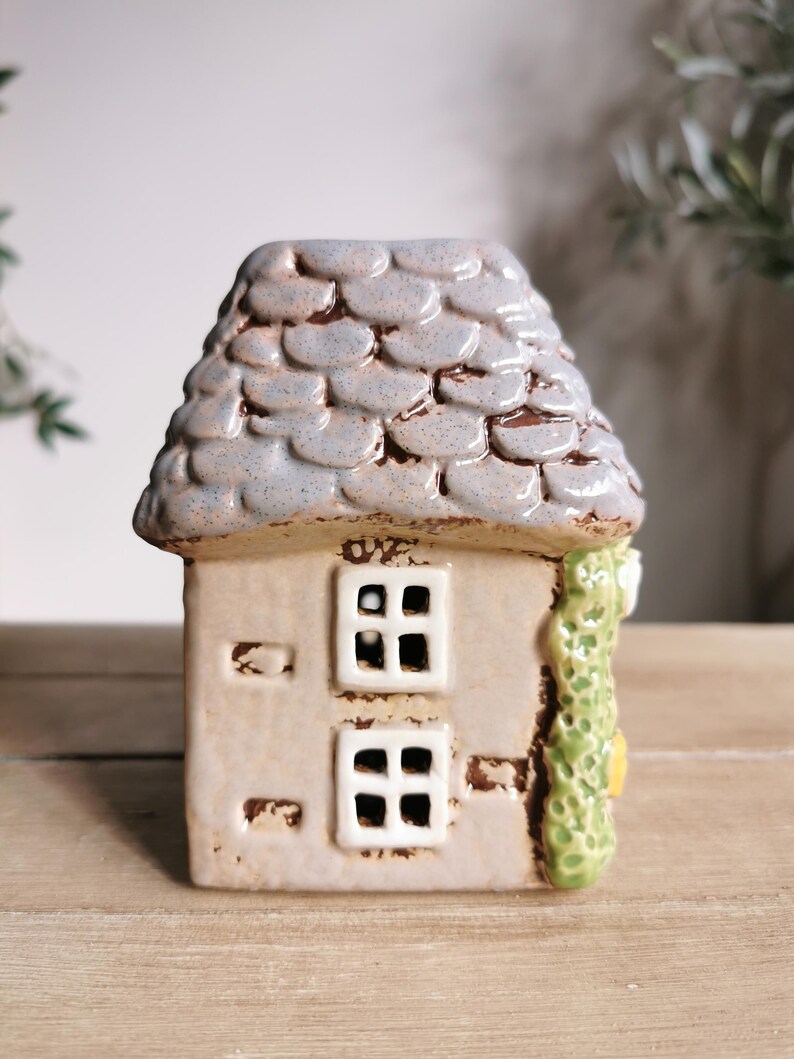 May include: A ceramic house figurine with a gray, textured roof and a beige body. The house has two square windows and a green vine detail on the side. The figurine is on a wooden surface.