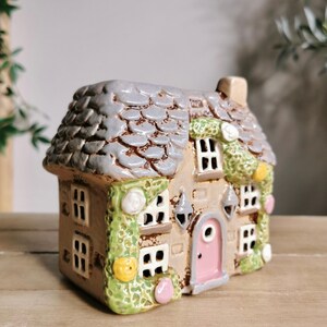 May include: A ceramic miniature house with a gray shingled roof, tan walls, and a pink door. The house has multiple windows, decorative green vines, and small, colorful accents. The overall design is whimsical and detailed.