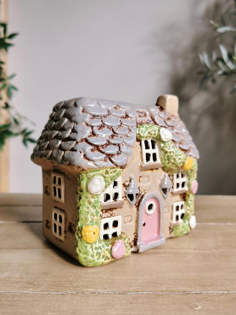 May include: A ceramic miniature house with a gray shingled roof, tan walls, and a pink door. The house has multiple windows, decorative green vines, and small, colorful accents. The overall design is whimsical and detailed.