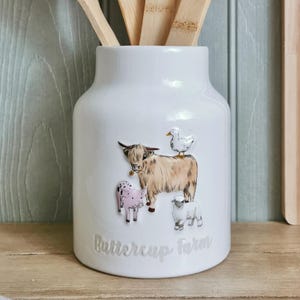 May include: A white ceramic utensil pot decorated with farm animals, including a Highland cow, pig, sheep, and duck. The words "Buttercup Farm" are written on the front. Wooden spoons are visible inside the pot.