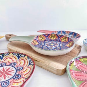 May include: Set of colourful ceramic spoon rests with intricate geometric and floral patterns. The rests are shaped like spoons with a handle and a shallow bowl. They are displayed on a wooden chopping board.