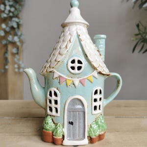 May include: A ceramic teapot in the shape of a house, painted in light blue with a white roof and chimney. The teapot features a door, windows, and a small bunting. Potted plants are at the base. A charming decorative item.