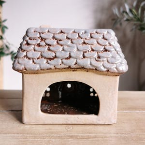 May include: A ceramic house-shaped candle holder. The roof is light blue with a textured, scale-like pattern, and the body is a cream color. The house has a large arched opening and small circular openings.