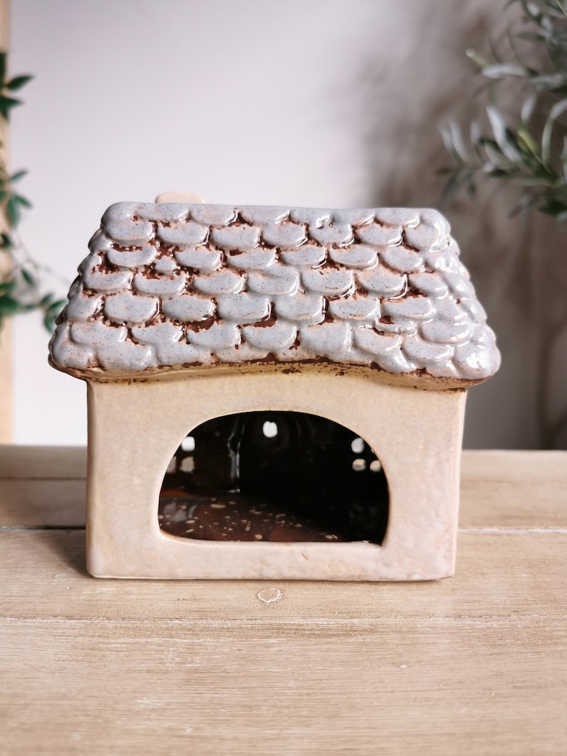 May include: A ceramic house-shaped candle holder. The roof is light blue with a textured, scale-like pattern, and the body is a cream color. The house has a large arched opening and small circular openings.