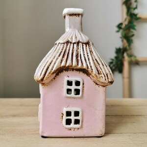 May include: A pink ceramic house with a thatched roof and two windows. The roof is tan with brown accents, and the windows are white. The house has a chimney and sits on a wooden surface. The house is a decorative item.