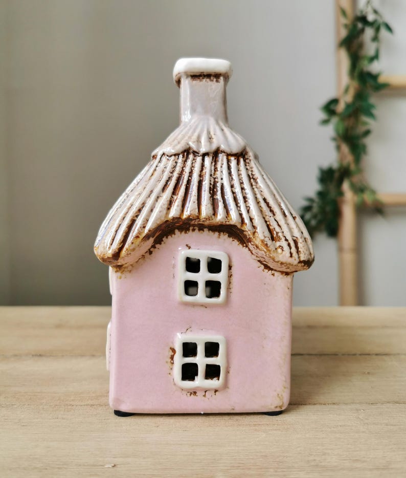 May include: A pink ceramic house with a thatched roof and two windows. The roof is tan with brown accents, and the windows are white. The house has a chimney and sits on a wooden surface. The house is a decorative item.