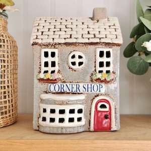 May include: A ceramic Corner Shop ornament with a grey exterior, white trim, and a red door. The roof is textured to resemble bricks. The shop has two windows with flower boxes and a round window. The sign above the window reads "CORNER SHOP".