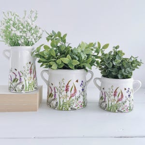 May include: Three white ceramic planters with floral designs featuring pink, purple, and white flowers and green leaves. The planters have two handles on each side and are filled with green plants.
