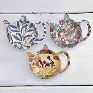 May include: Three ceramic tea bag tidies shaped like teapots. One has a blue and white leaf design, another a floral and bird pattern, and the third depicts a farm scene with dogs and a tractor. Each is a unique design.