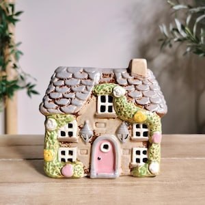 May include: A ceramic cottage with a gray shingled roof, beige walls, and a pink door. The windows are white, and the cottage is adorned with green and yellow floral accents. Two silver sconces flank the door.