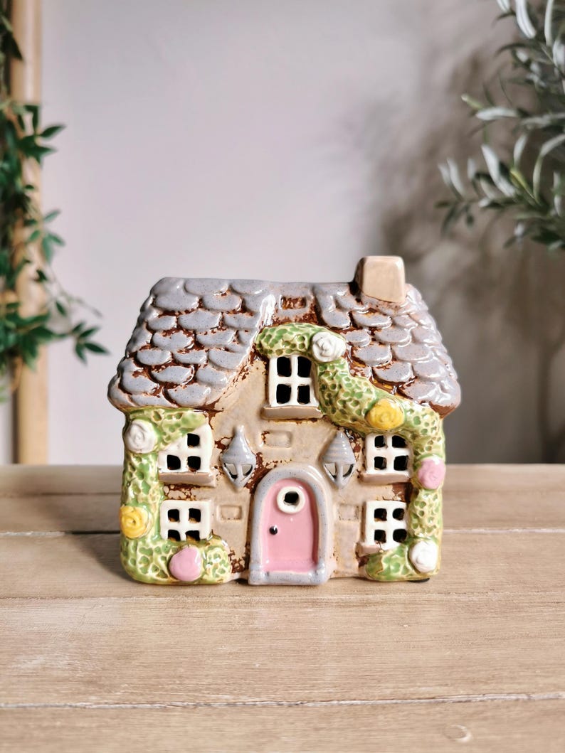 May include: A ceramic cottage with a gray shingled roof, beige walls, and a pink door. The windows are white, and the cottage is adorned with green and yellow floral accents. Two silver sconces flank the door.