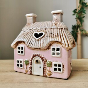May include: A pink ceramic cottage with a thatched roof and heart-shaped window. The cottage has four square windows, a white door, and two chimneys. Decorative potted trees flank the door. The cottage is a decorative item.