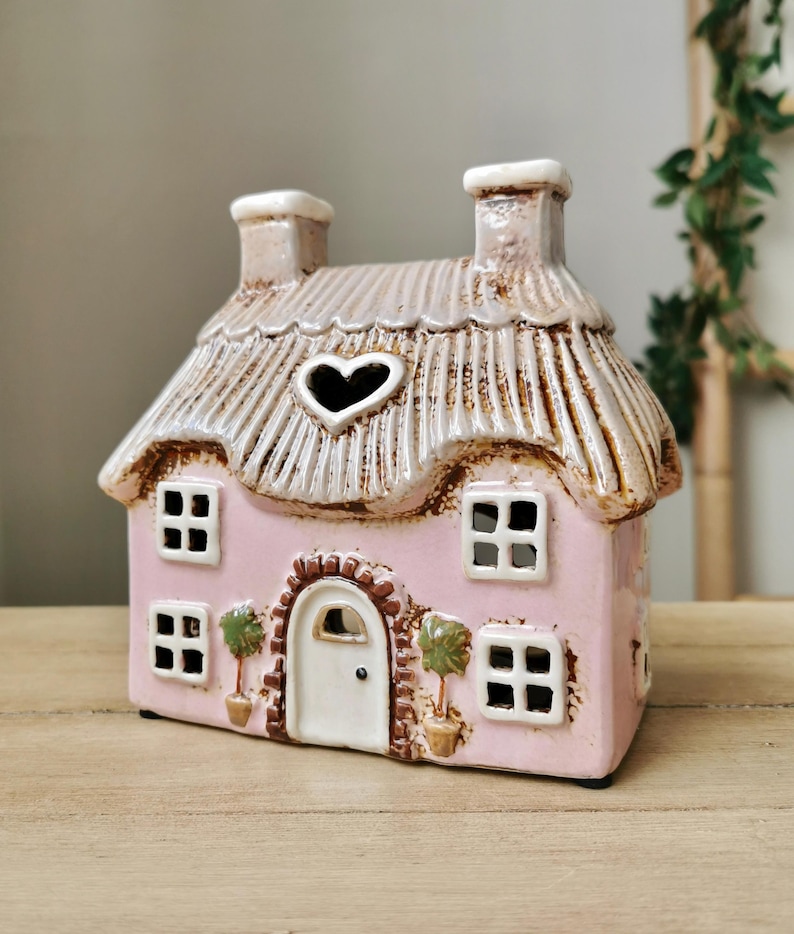May include: A pink ceramic cottage with a thatched roof and heart-shaped window. The cottage has four square windows, a white door, and two chimneys. Decorative potted trees flank the door. The cottage is a decorative item.