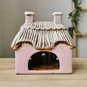 May include: A pink ceramic cottage with a thatched roof and two chimneys. The cottage has a large arched opening and is set on a wooden surface. The roof is white with brown accents.