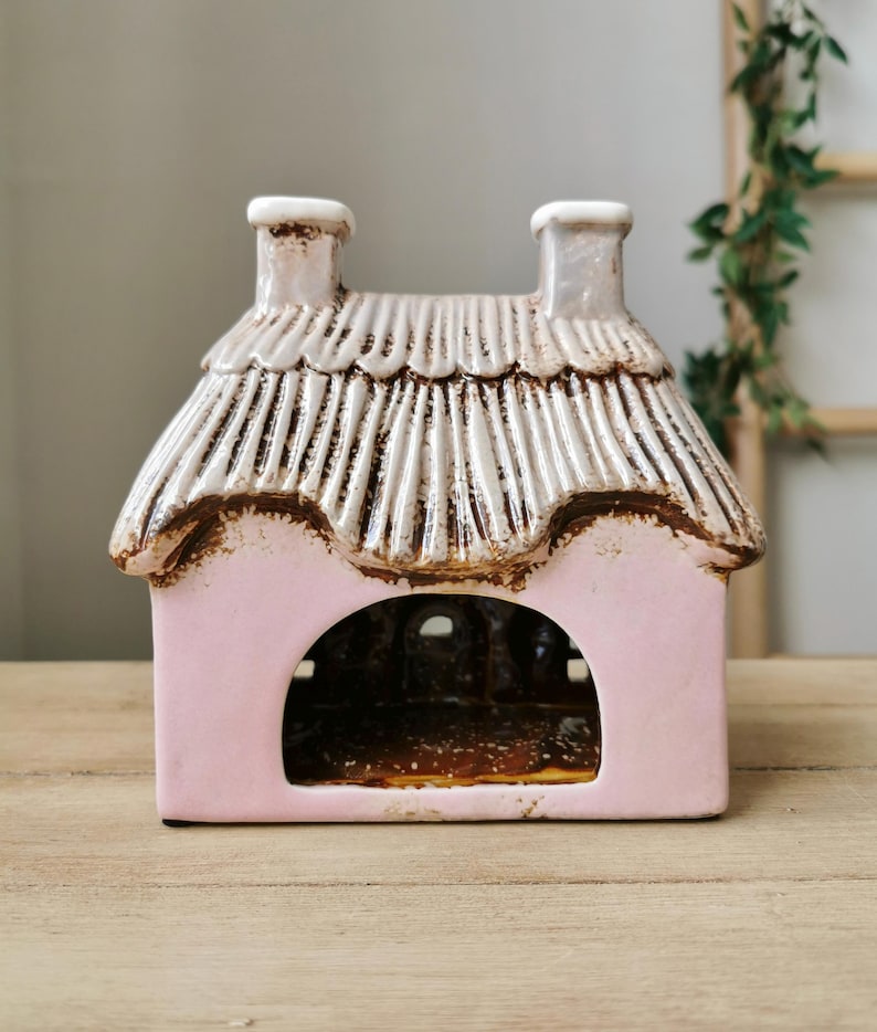 May include: A pink ceramic cottage with a thatched roof and two chimneys. The cottage has a large arched opening and is set on a wooden surface. The roof is white with brown accents.