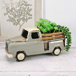 May include: A grey ceramic pick-up truck planter with a wooden bed, filled with green succulent plants.