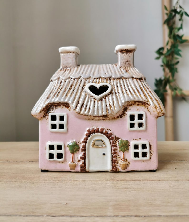 May include: A pink ceramic house-shaped candle holder with a heart-shaped window. The roof is tan and textured, with two chimneys. The house has four square windows and a white door. Two small potted trees flank the door.
