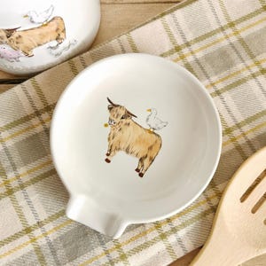 May include: White ceramic spoon rest featuring a Highland cow and duck illustration. The spoon rest has a curved shape with a small lip for resting utensils. The cow is brown with horns and a yellow flower in its mouth.