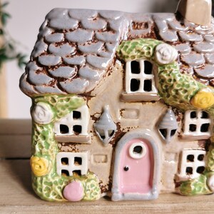 May include: A ceramic cottage with a gray shingled roof, a pink door, and white-framed windows. Green vines with white and yellow flowers adorn the exterior. The cottage has a chimney and decorative hanging lanterns.