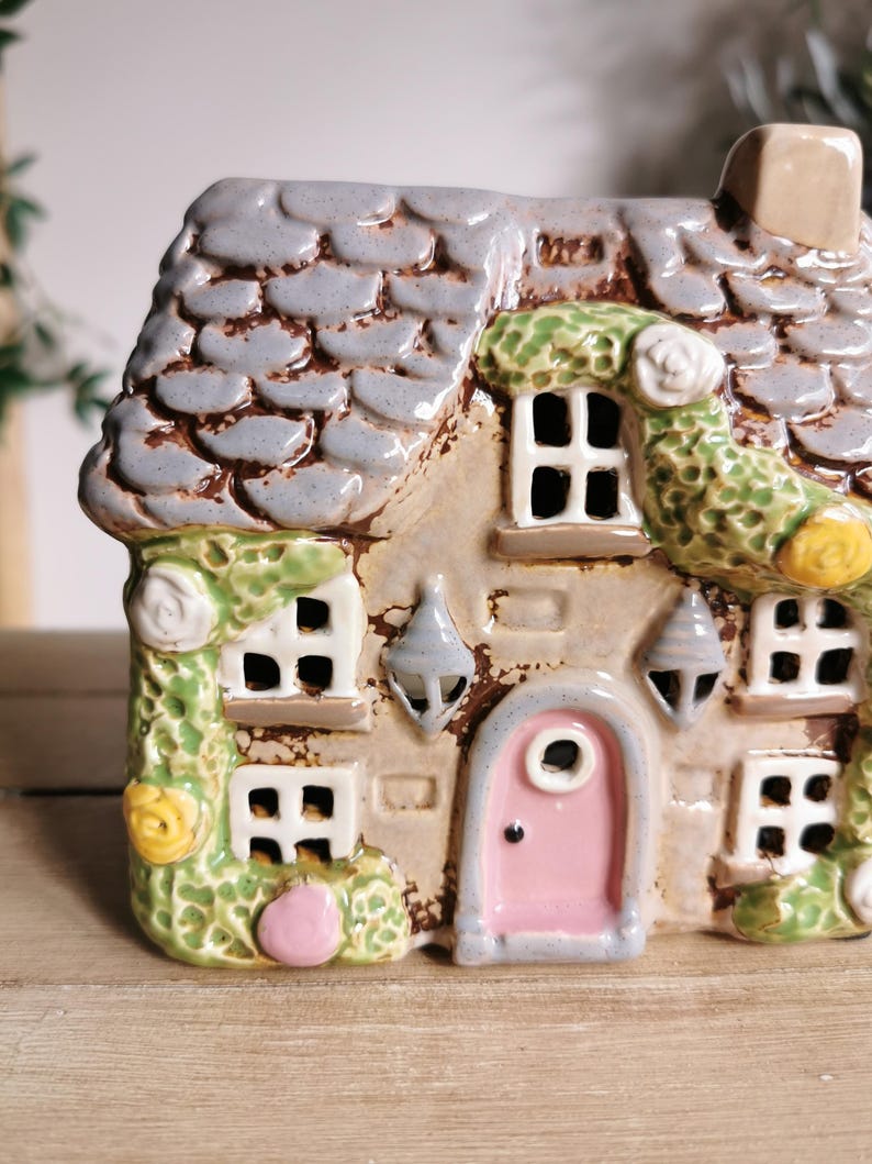 May include: A ceramic cottage with a gray shingled roof, a pink door, and white-framed windows. Green vines with white and yellow flowers adorn the exterior. The cottage has a chimney and decorative hanging lanterns.