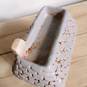 May include: A small, rectangular ceramic planter with a textured, basket-weave design. The planter is primarily light blue with brown accents and a small, off-white cube attached to the side. The interior is a darker shade of blue.
