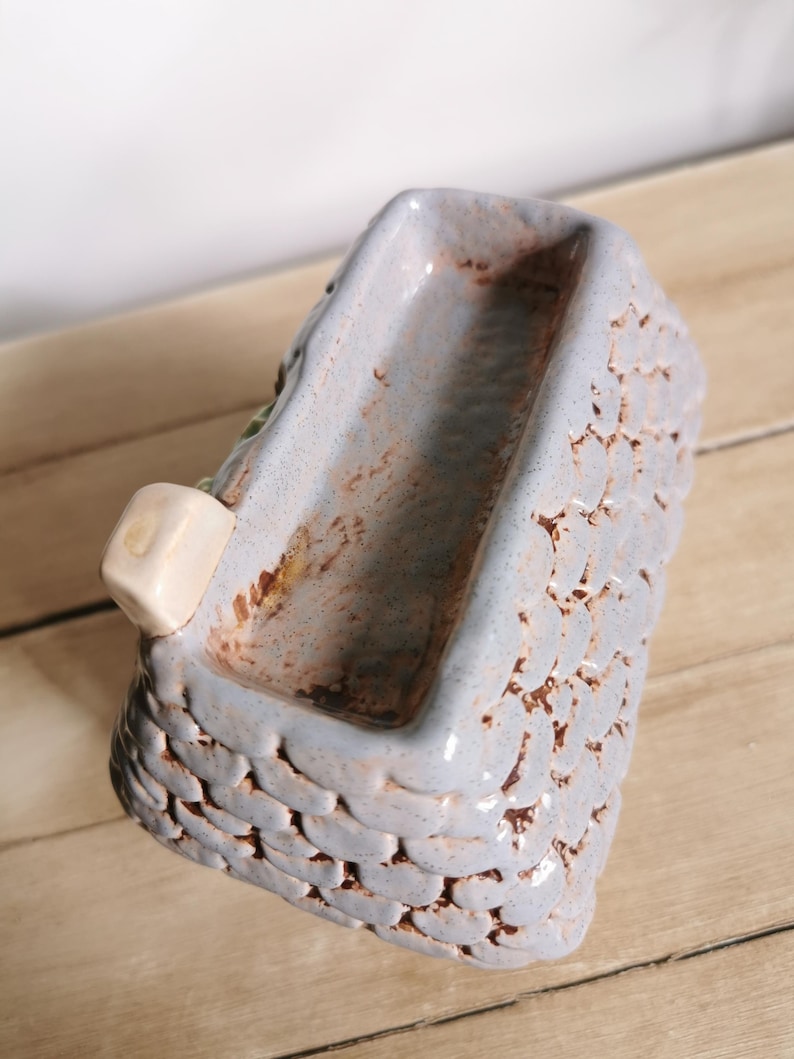 May include: A small, rectangular ceramic planter with a textured, basket-weave design. The planter is primarily light blue with brown accents and a small, off-white cube attached to the side. The interior is a darker shade of blue.