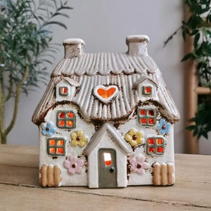 May include: A ceramic cottage-shaped candle holder with a thatched roof, chimneys, and flower accents. The windows glow with an orange light, and a heart shape is on the roof. The cottage is white with brown and pastel-coloured details.
