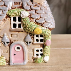 May include: A ceramic cottage figurine with a gray tiled roof, a pink door, and green vines with white and yellow flowers. The cottage has small windows and decorative hanging lights. The overall color scheme is neutral with pastel accents.