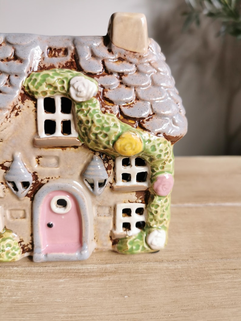 May include: A ceramic cottage figurine with a gray tiled roof, a pink door, and green vines with white and yellow flowers. The cottage has small windows and decorative hanging lights. The overall color scheme is neutral with pastel accents.