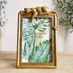 May include: A gold-coloured rectangular photo frame with a textured border, featuring a decorative leopard resting on top. The frame holds a print with tropical leaves and the text "Photo paper 10 x 15 cm".
