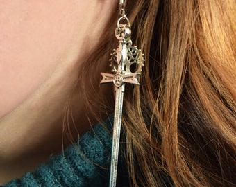 May include: Silver sword earrings with a detailed design. The sword has a crown at the top and a cross on the blade.