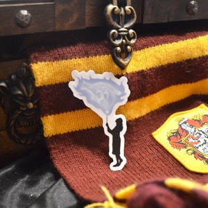 May include: A white sticker with a silhouette of a person holding a blue balloon. The sticker is on a brown and gold striped scarf.