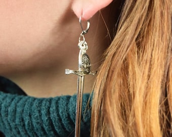 May include: Silver sword earrings with a detailed design. The earrings are hanging from a person's ear.