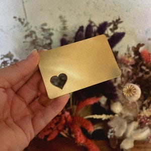 Op de afbeelding: Een goudkleurige rechthoekige metalen kaart met een hartvormige uitsparing. De kaart wordt in een hand gehouden, met een vage achtergrond van gedroogde bloemen in rood, paars en beige.