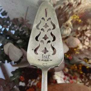 Puede incluir: Un servidor de pastel plateado con un diseño calado decorativo. El servidor tiene las palabras "fait avec" y un símbolo de corazón grabado en el mango. El fondo presenta flores secas.