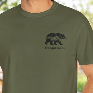 May include: Olive green t-shirt featuring a black graphic of a bear with a forest scene and a crescent moon. The text "Fappa Bear" is printed below the graphic. The shirt has short sleeves and a crew neck.
