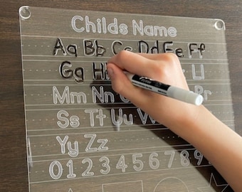 May include: A clear plastic dry-erase board with white outlines of the alphabet, numbers 0-9, and basic shapes. The board has a hole in the top corner for hanging.