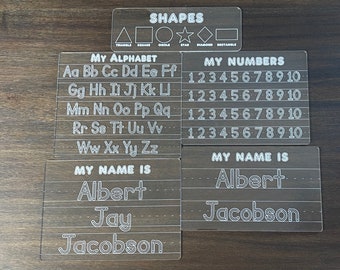 May include: Four clear plastic dry-erase boards with white outlines. The top board has the word "SHAPES" and outlines of a triangle, square, circle, star, diamond, and rectangle. The second board has the words "MY ALPHABET" and outlines of the letters of the alphabet. The third board has the words "MY NUMBERS" and outlines of the numbers 1 through 10. The fourth board has the words "MY NAME IS" and outlines for the name "Albert Jacobson".