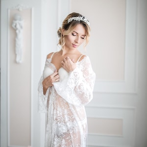 May include: A woman wearing a white lace robe and a matching bra. She is also wearing a silver leaf-shaped headband. The robe has long sleeves and a floral lace pattern. The background is a light-colored room with decorative wall panels.