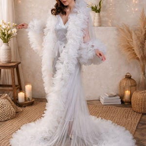 May include: A white, floor-length robe with layered ruffles and a long train. The robe has a V-neck and long sleeves with ruffled cuffs. The image is set in a room with soft lighting, candles, and decorative plants.