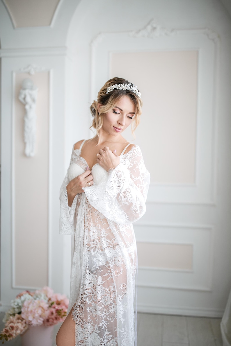May include: A woman wearing a white lace robe and a matching bra. She is also wearing a silver leaf-shaped headband. The robe has long sleeves and a floral lace pattern. The background is a light-colored room with decorative wall panels.
