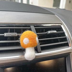 May include: A felt mushroom car air freshener in orange and white. The mushroom is hanging from the air vent of a car.