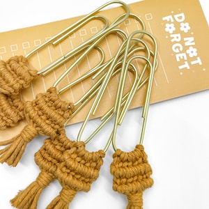 May include: Gold-colored paper clips with a woven, mustard-colored tassel detail. The paper clips are arranged on a tan notepad with the words "DON'T FORGET" printed in white.
