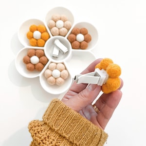 May include: A set of six felt flower car air fresheners in various colors, including orange, brown, and white. The flowers are arranged in a white flower-shaped tray. One flower is being held in a hand, showing the clip on the back.