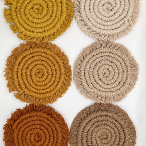 May include: Six round crocheted coasters with fringe. The coasters are in shades of brown, beige, and yellow.